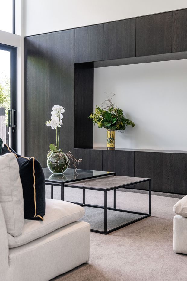 A refined contemporary living room with a full-height dark timber built-in entertainment and display wall, featuring open white niches and lower cabinetry. A pair of slim black-framed coffee tables sits on a pale carpet between cream upholstered sofas. Natural light enters through a tall glazed door or window, while orchids and a floral arrangement add organic detail.