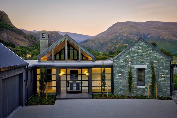 Exterior view of a contemporary mountain residence at dusk, composed of dark metal-clad volumes, a glazed gable-roofed central entry pavilion, and a prominent green-grey stone gable wall. The front facade includes large windows, warm interior lighting, a covered entry, landscaping, and broad paved access areas, with dramatic alpine mountains behind.