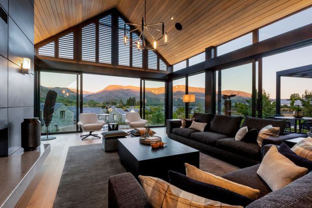 A sophisticated contemporary living room with a large dark sectional sofa arranged around a low black coffee table and textured rug. Floor-to-ceiling glazing opens to mountain and suburban views, while a dramatic slatted timber ceiling, exposed dark structural beams, clerestory louvres, and a sculptural pendant light create a warm, architectural interior.