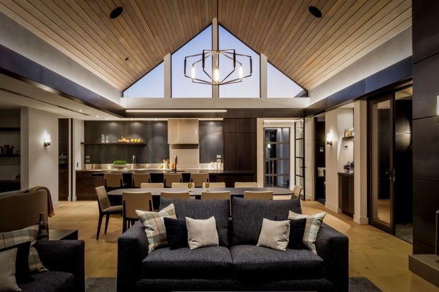 Open-plan contemporary living, dining and kitchen interior with a dark upholstered sofa in the foreground, dining table and chairs centrally positioned, and a modern kitchen with dark cabinetry and a light island at the rear. The room has a dramatic pitched timber-lined ceiling, clerestory glazing, exposed dark structural beams, recessed lighting, and a geometric pendant light.