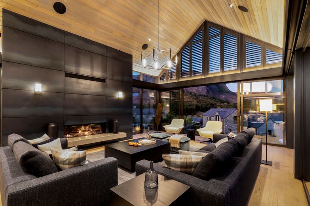 A sophisticated double-height living room with a dramatic dark feature wall, built-in fireplace, expansive glazing, and a warm timber-lined vaulted ceiling. Multiple charcoal sofas and cream lounge chairs surround a low dark coffee table, while high-level timber louvres and exposed structural beams add architectural interest. The glazing looks toward a mountainous landscape and adjacent pitched-roof buildings.