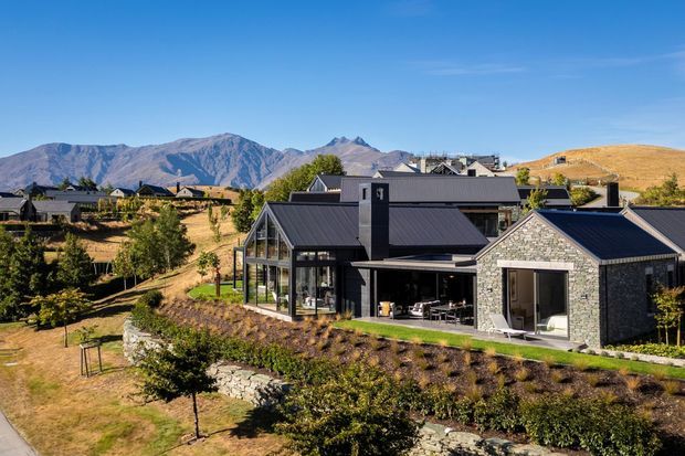 Exterior view of a contemporary single-storey residence set within a landscaped, sloping site. The home has dark metal pitched roofs, extensive glazed living areas, and a prominent gabled wing clad in grey natural stone. Dry grassland, stone retaining walls, and rugged mountains form the surrounding setting.