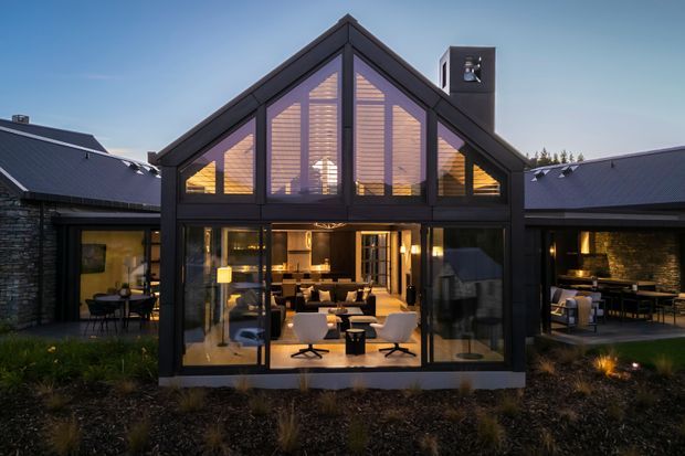 Twilight exterior view of a contemporary gable-roof house with a central double-height glazed living space. Dark metal-framed glazing and cladding contrast with stone side wings, while warm interior lighting reveals a living room and dining area. The home is surrounded by landscaped planting and connected low-pitched roof volumes.