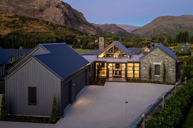 Twilight exterior view of a contemporary mountain residence, with multiple steep gabled roof volumes, dark vertical cladding, a stone-faced wing, expansive illuminated glazing, and a broad concrete driveway. Rugged mountainous terrain and mature landscaping form the backdrop.