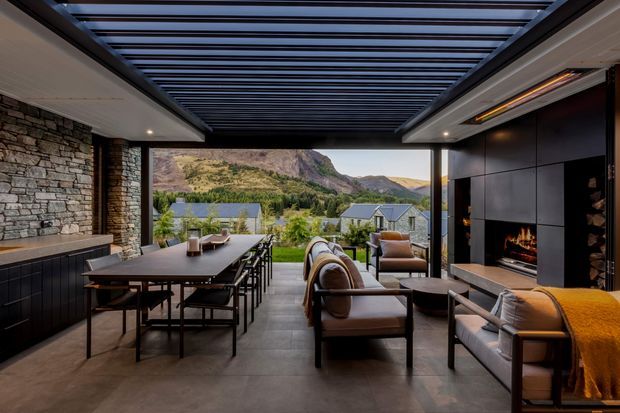 A sophisticated covered outdoor entertaining terrace overlooking a dramatic mountain landscape. The space features a long dining table, lounge seating, a built-in linear fireplace within dark wall panelling, a stone feature wall with integrated cabinetry, a concrete-look floor, and a dark overhead louvred pergola.