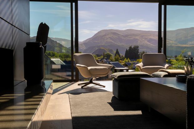 A sophisticated contemporary living room with floor-to-ceiling glazing overlooking a dramatic mountainous landscape. The room features sculptural cream lounge chairs, a dark low-profile ottoman or coffee table, a large dark rug, timber flooring, dark wall panels, and a sleek black built-in media unit. Strong natural light and expansive views create a refined indoor-outdoor connection.