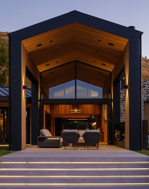 A dramatic dusk exterior view of a modern multi-level residence with a tall, open gable-like timber-lined volume framing a glazed living area. The dark charcoal facade contrasts with warm timber soffits and wall lining, while broad illuminated steps lead up to an outdoor seating area. Large glazing reveals the interior, and a stone-clad side wall and sloping landscaped site are visible.