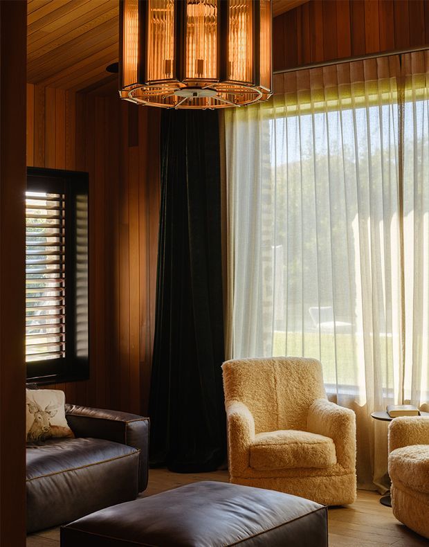A warm, richly toned living room with dark timber wall and ceiling panelling, a large sheer-curtained window, dark leather seating and ottoman, and plush cream boucle armchairs. A sculptural brass-toned pendant light hangs prominently overhead, while a black curtain and louvred window add contrast.