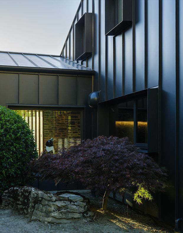 A moody architectural exterior view of a contemporary dark-clad building, showing a tall metal-clad facade with vertical seams, projecting window surrounds, and a low adjoining wing with a standing-seam metal roof. Glazed openings reveal a softly lit interior, while layered stone landscaping and mature shrubs frame the entrance courtyard.