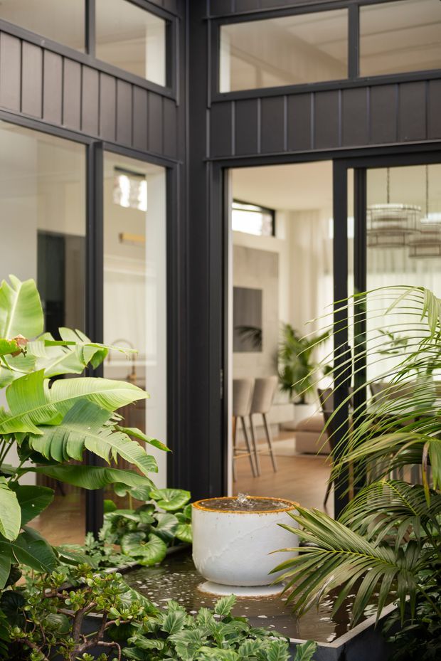 A lush indoor courtyard or atrium viewed from within, featuring dense tropical planting, a shallow water feature with a round white planter or fountain, and dark metal-framed glazing. The glazing reveals a bright contemporary dining area beyond, with warm timber flooring, neutral walls, and a sculptural hanging light. The composition contrasts black framed windows and cladding with abundant greenery and soft natural light.