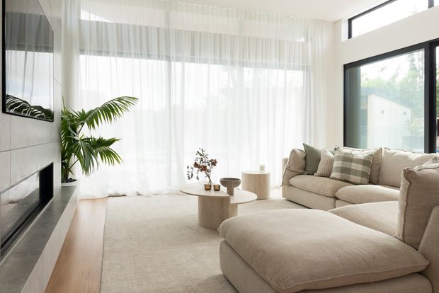 Bright contemporary living room with a large cream sectional sofa and ottoman, layered neutral textiles, a low round timber coffee table, a linear fireplace with dark surround, sheer full-height curtains, indoor palm, and expansive black-framed glazing. The room has a calm, refined, natural palette and abundant daylight.