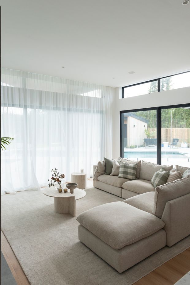 Bright contemporary living room with a large cream sectional sofa, oversized ottoman, pale textured rug, round stone-look coffee table, sheer full-height curtains, and expansive black-framed glazing. The glazing overlooks a swimming pool and landscaped outdoor area, while the interior uses a soft neutral palette and abundant natural light.
