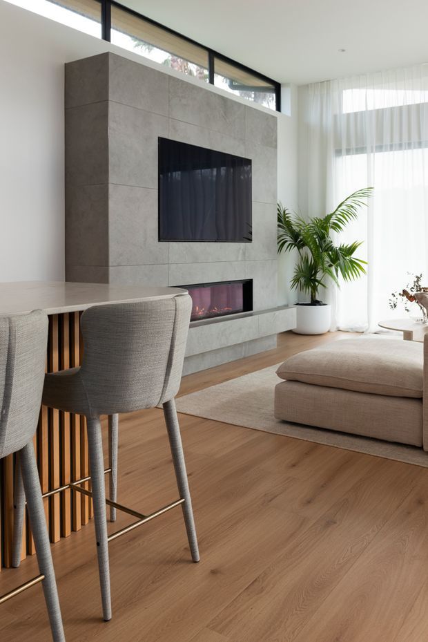 Bright contemporary open-plan living and dining area with a warm timber floor, a large concrete-look tiled media wall incorporating a wall-mounted television and linear fireplace, and full-height sheer-curtained glazing. A neutral upholstered sofa and ottoman sit beside a light dining or kitchen island with upholstered bar stools and timber slat detailing. Indoor palms and soft beige furnishings add warmth to the clean, refined interior.