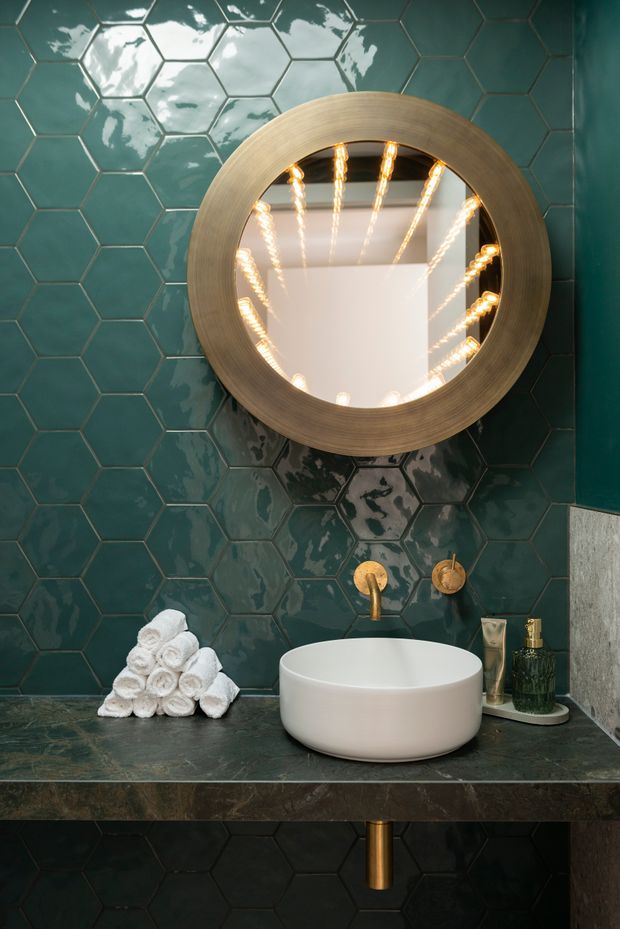 A moody, luxurious bathroom vanity vignette featuring glossy dark teal hexagonal wall tiles, a round brushed brass-framed mirror with reflected strip lighting, a white vessel basin on a dark stone countertop, and brass wall-mounted tapware. Rolled white towels and toiletries sit beside the basin, with dark built-in cabinetry below.