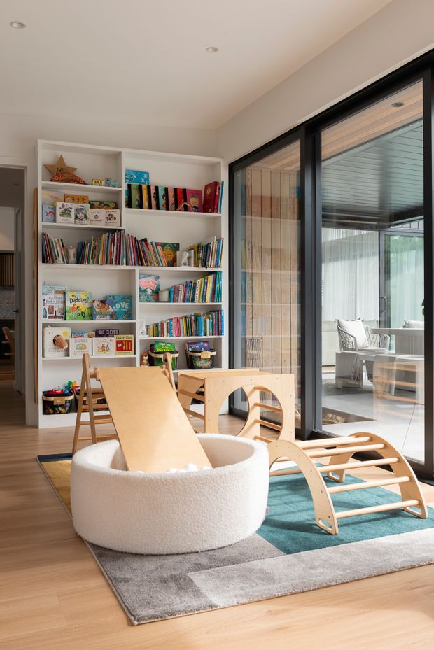Bright, contemporary children's playroom with a large white built-in bookshelf filled with colourful books and toys, light timber play furniture, a soft round boucle play pit, and a grey-and-turquoise area rug. Full-height black-framed glazing opens visually to a covered outdoor space, while pale timber flooring and recessed ceiling lights create an airy, warm setting.