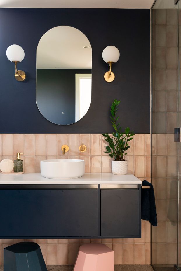 A stylish contemporary bathroom vanity featuring dark navy wall panelling, warm blush-pink square wall tiles, a white vessel basin on a pale benchtop, dark built-in cabinetry, brass wall sconces and tapware, and a rounded vertical mirror. A glass shower enclosure is visible at right, with a small plant and colourful stools adding decorative accents.