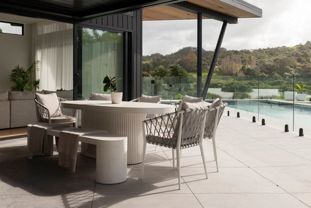 A contemporary covered outdoor dining terrace beside a swimming pool, with a round fluted dining table, woven dining chairs, stools, and a built-in lounge area. Large glazed openings connect the terrace to the interior, while dark vertical cladding, timber-lined soffits, slim black structural posts, and frameless glass pool balustrading frame views toward a wooded landscape.