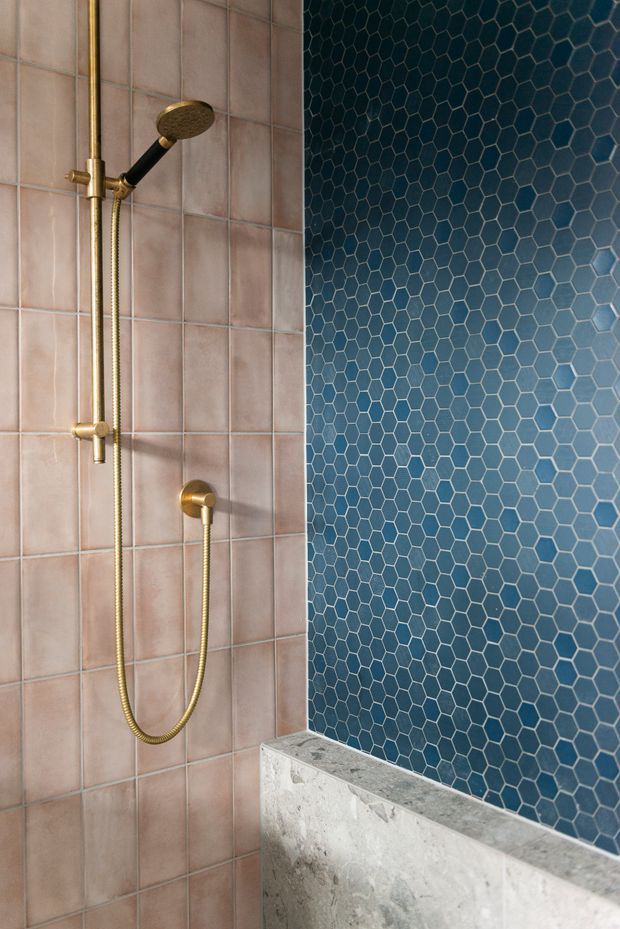 Close-up of a contemporary shower enclosure featuring brushed brass shower fittings, blush-pink square wall tiles, a deep blue hexagonal tile feature wall, and a pale textured stone or concrete ledge.