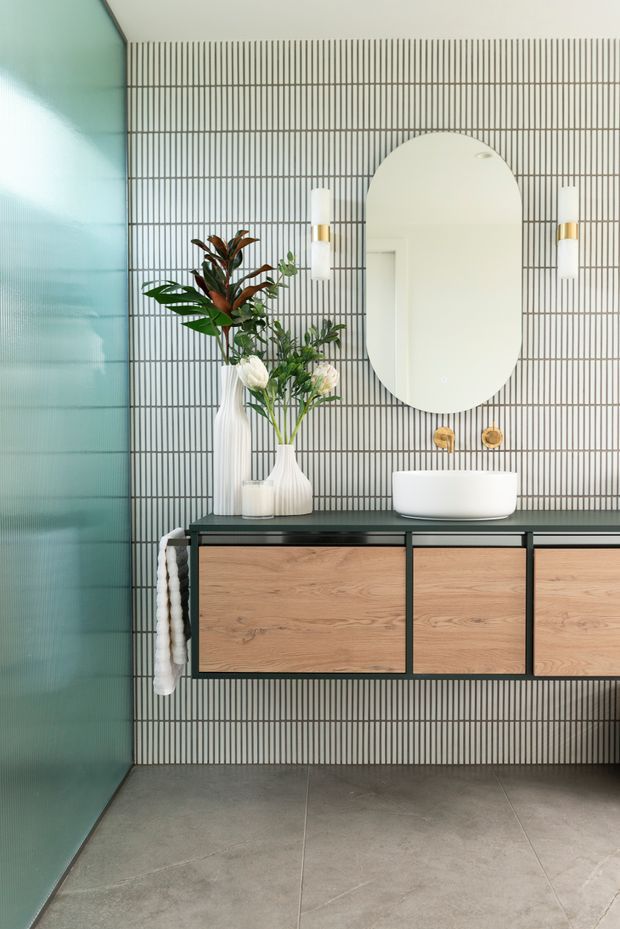 A contemporary bathroom vanity area with a wall-mounted timber-and-black vanity, white vessel basin, oval mirror, brass wall-mounted tapware, and vertical striped grey wall tiles. A ribbed translucent green glass wall or door is visible at left, with decorative vases and greenery on the vanity. Large grey floor tiles complete the space.