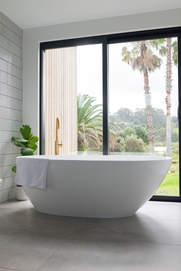 Bright contemporary bathroom featuring a freestanding white bathtub positioned beside large black-framed glazed doors, with a brass floor-mounted tap, grey tiled floor, vertically tiled wall, and views of palm trees and landscaped grounds outside.