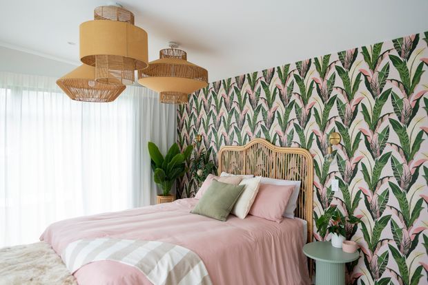 A bright, playful bedroom featuring a rattan bedhead, pink bedding, layered cushions, botanical feature wallpaper, woven pendant lights, sheer curtains, and a small sage-green bedside table. Indoor plants add a natural accent to the room.