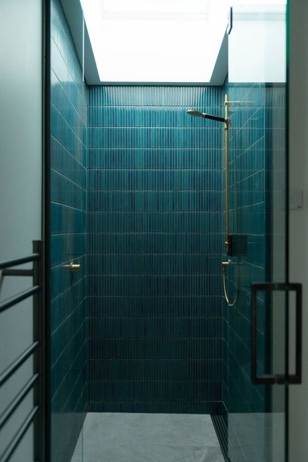 A narrow, contemporary walk-in shower enclosure viewed through a glass door. The shower walls are fully clad in deep teal-blue vertically ribbed ceramic tiles, with brass/gold-toned shower fittings and a pale grey tiled floor. A bright skylight or ceiling opening illuminates the compact space, while dark-framed glass and balustrade elements appear at the edges.