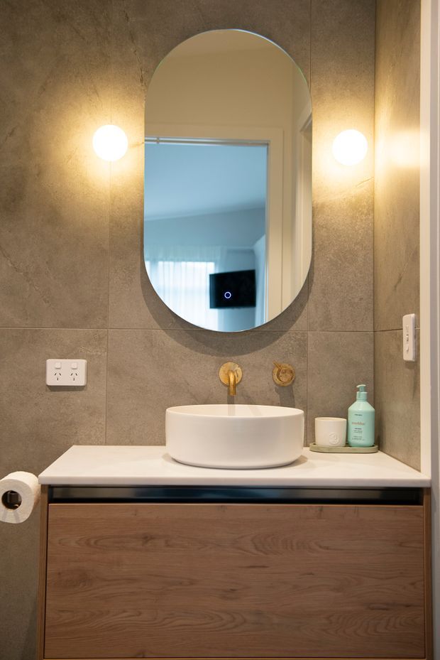A compact contemporary bathroom vanity with a round vessel basin on a white benchtop, wall-mounted gold-toned tapware, a large arched mirror, warm globe wall lights, grey stone-look wall tiles, and timber-grain drawer cabinetry.