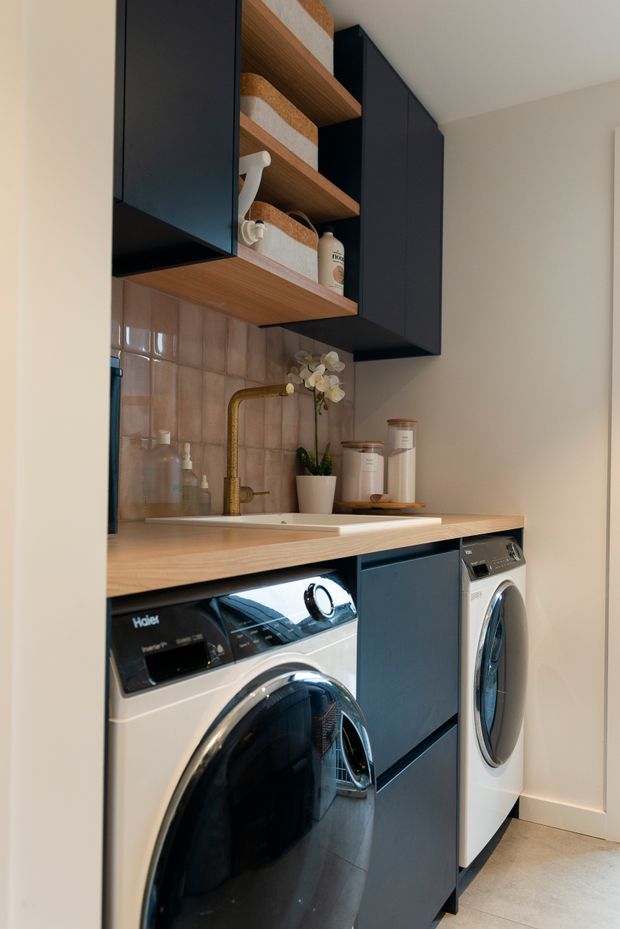 A compact, well-organised laundry room with stacked front-loading washer and dryer units beneath a light timber benchtop. Dark navy built-in cabinetry provides concealed storage, while open timber shelves display baskets and containers. A brass-toned wall-mounted tap, pale pink-beige glazed tile splashback, sink, and small decorative plant add warmth and detail. The room has a clean, contemporary fitted-out appearance.