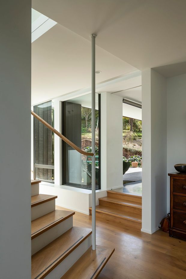 A bright contemporary stairway and landing with warm timber flooring and open-riser stairs, a slim central metal support, and a curved timber handrail. White walls and ceilings frame large glazed openings overlooking a landscaped outdoor area, with natural light entering through a skylight and full-height windows. A dark timber chest sits at the edge of the landing.