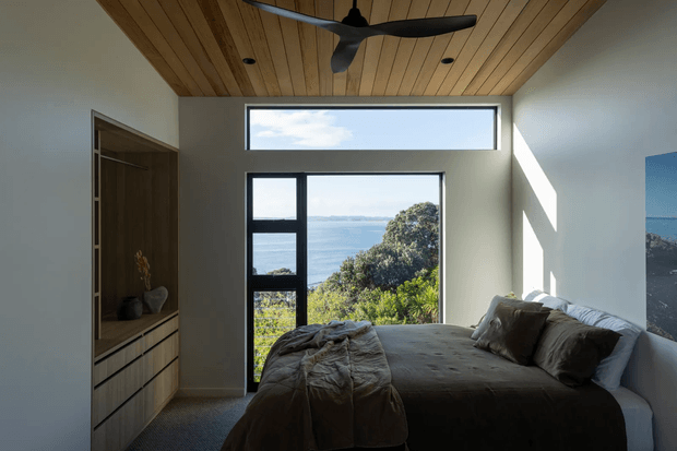 A compact, contemporary bedroom with a low platform bed facing large windows overlooking a blue coastal or ocean landscape. The room has white plaster walls, a warm timber-lined ceiling with a black ceiling fan, dark-framed glazing including a high clerestory window, and built-in timber cabinetry with drawers and open shelving. Strong sunlight creates diagonal shadows across the wall, giving the space a calm, natural atmosphere.