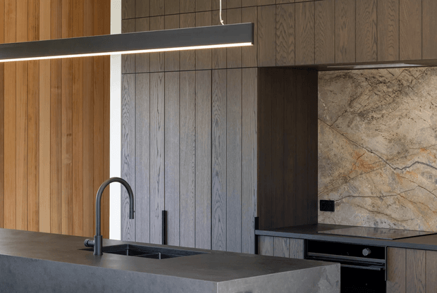 Close-up view of a refined contemporary kitchen featuring a large dark stone island with integrated sink and curved black tap, vertical timber wall panelling, dark wood cabinetry, a dramatic natural-stone splashback, and a slim linear pendant light.