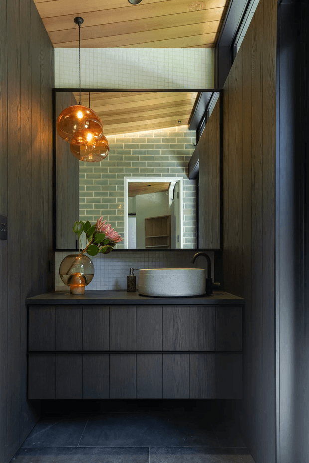 A compact, moody bathroom vanity with a floating dark timber-grain cabinet, dark stone benchtop, and round light-coloured vessel basin. A large black-framed mirror reflects a green glazed-brick wall, timber-lined ceiling, and roof glazing. Warm amber globe pendant lights and a small vase provide contrast against the dark joinery and floor.