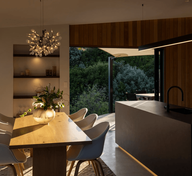 A warm, contemporary open-plan kitchen and dining area with a large timber dining table, sculptural pendant light, integrated shelving, and a dark kitchen island with a black gooseneck tap. Full-height glazing opens toward a leafy outdoor setting, while timber wall panelling and concealed linear lighting add depth and refinement.