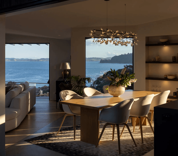 A warm, refined open-plan dining and living area overlooking a blue coastal inlet through large windows. The room features a substantial timber dining table with upholstered chairs, a sculptural branching pendant light, built-in illuminated shelving, indoor plants, a neutral sofa, and dark timber or stone flooring. A rocky shoreline and wooded headland are visible outside.