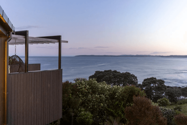 A coastal exterior view from a raised deck or balcony, with a dark vertical-clad privacy screen, pergola-like timber structure, and hanging egg chair in the foreground. The property overlooks dense coastal vegetation and a broad expanse of ocean toward a distant shoreline under a pale evening sky.