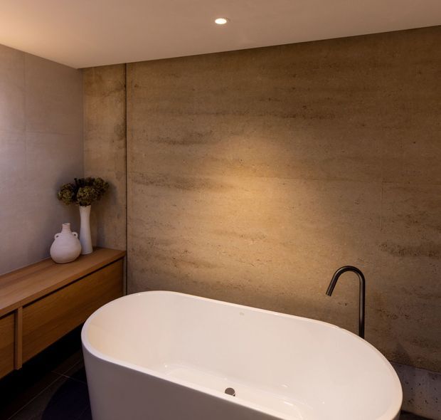 A warm, refined bathroom featuring a freestanding white bathtub in the foreground, a dark curved floor-mounted tap, and a large textured brown concrete or stone feature wall. A timber vanity with drawers and decorative ceramic vessels sits to the left, with recessed ceiling lighting creating a softly lit, spa-like atmosphere.