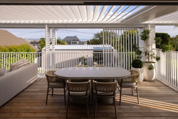 A bright covered outdoor terrace with a timber deck, oval dining table and woven dining chairs, accompanied by a light outdoor sofa. White vertical screening and railings frame suburban views, while a slatted pergola roof creates patterned sunlight and shade. Potted topiary plants add greenery to the refined entertaining space.