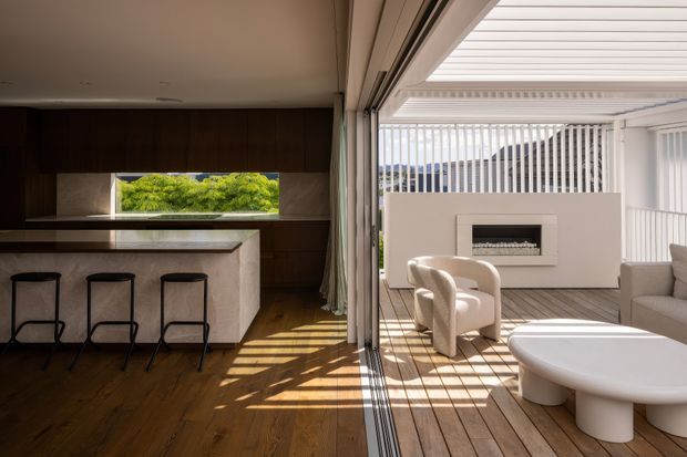 A refined open-plan interior opening onto a bright covered outdoor entertaining area. The room features dark timber wall panelling and flooring, a pale stone kitchen island with three bar stools, and a large sliding glazed opening. Outside, a timber deck includes sculptural lounge seating, a rounded white coffee table, and a minimalist built-in fireplace set against a slatted privacy screen. Strong sunlight creates linear shadows across the floor and deck.