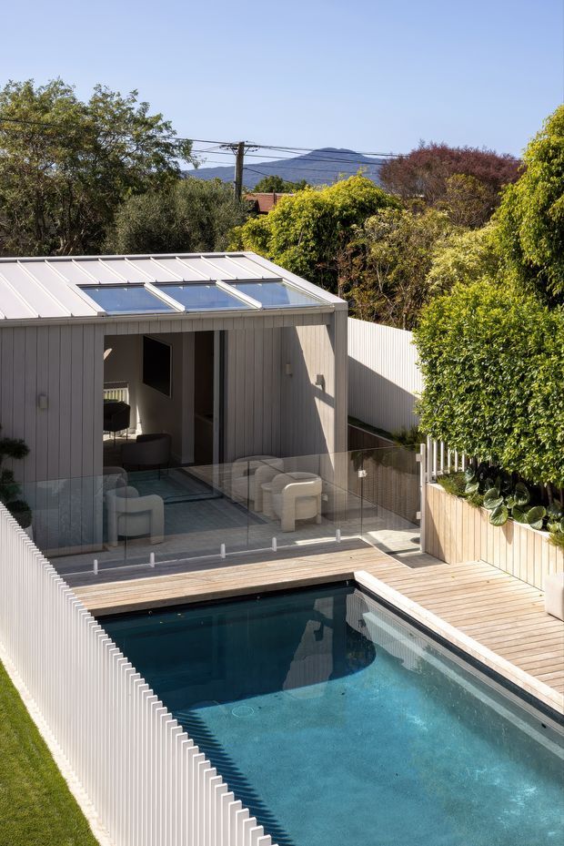 Elevated exterior view of a compact contemporary backyard pavilion beside a swimming pool. The pavilion has light vertical cladding, broad roof skylights, glazed walls and sliding doors, and a small furnished lounge opening onto a timber pool deck. White privacy fencing, planted raised beds, mature trees and distant hills surround the pool area.