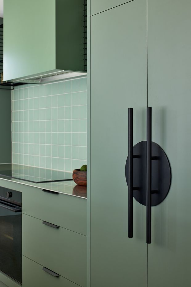 Close-up view of a contemporary kitchen with muted sage-green cabinetry, tall pantry doors, long matte-black vertical handles, a matching dark benchtop, pale green square-tile splashback, and integrated dark appliances. The composition focuses on the fixed joinery and hardware, with a small timber bowl on the counter.
