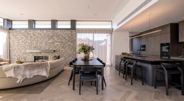 Open-plan contemporary living, dining, and kitchen area with a double-height feel, expansive clerestory glazing, a full-height stacked-stone fireplace wall, dark timber kitchen cabinetry, stone island benchtop, and light herringbone timber flooring. The room is furnished with a dining table, island stools, and a curved sectional sofa, creating a refined yet inviting interior.