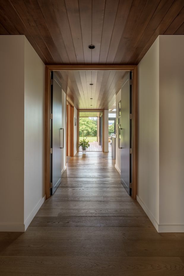 A warm, elongated interior hallway with wide grey-brown timber flooring and a matching timber-lined ceiling. White plaster walls frame a series of open glazed and timber doors, leading toward a bright exterior deck and landscaped view. Minimal recessed and pendant lighting, dark door hardware, and strong axial perspective create a calm, refined circulation space.
