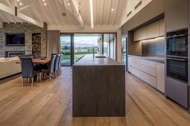 A spacious open-plan kitchen and dining area with a large dark stone island, integrated timber-toned cabinetry, built-in black appliances, and a dining table beside a rustic stone fireplace wall. The room has warm timber flooring, exposed white roof beams, recessed lighting, and full-height glazing opening toward a landscaped view.