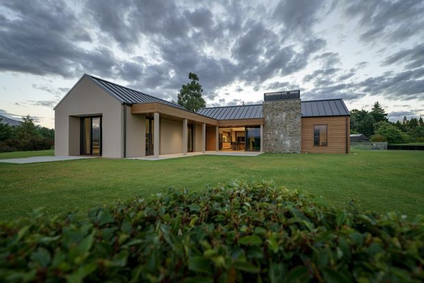 A single-storey contemporary home set within a broad landscaped lawn, viewed from the front. The house has multiple pitched roof forms with dark standing-seam metal roofing, pale rendered walls, warm timber cladding, large glazed openings, and a prominent rough stone-clad chimney or feature wall. A covered recessed entry and courtyard-like central opening connect the wings of the house. The scene is photographed under a dramatic overcast sky with hedging in the foreground.