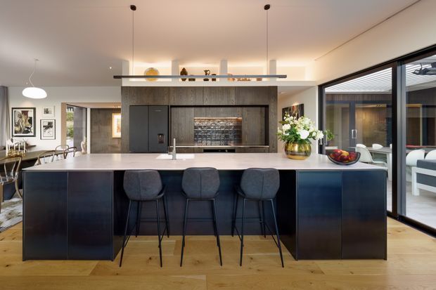 Contemporary open-plan kitchen centred on a large island with dark integrated cabinetry, a pale stone benchtop and seating for four. The room features timber flooring, a dark textured feature wall and fireplace or media recess, a linear pendant light, recessed display shelving, and extensive glazing opening toward an outdoor area. A dining zone and living furnishings are visible beyond the island.