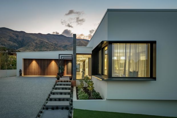 Contemporary single-storey residence with a crisp white and dark-framed facade, flat rooflines, deep-set glazing and a timber-toned garage door. A gravel driveway and landscaped stepped entry lead to the illuminated entrance. Mountainous terrain is visible behind the home.