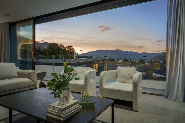 Sophisticated living room with a dark rectangular coffee table, upholstered lounge chairs, a large neutral sofa, floor-to-ceiling glazing, and full-height curtains. The room opens to expansive mountain and valley views at dusk, creating a refined, calm atmosphere with a neutral palette and strong indoor-outdoor connection.