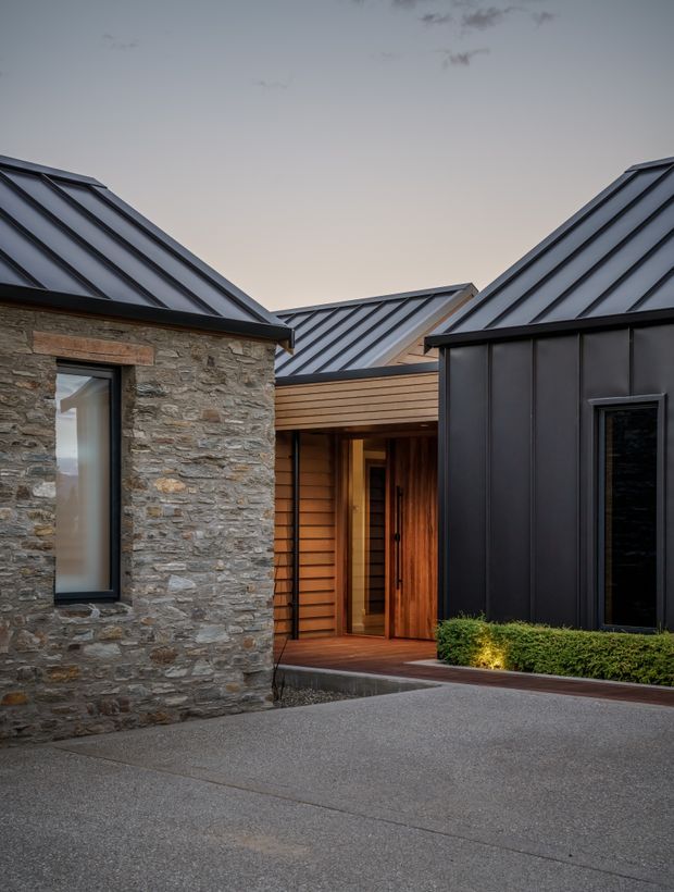 Exterior architectural photograph of a contemporary multi-wing residence arranged around an entry courtyard. The building combines rough natural stone walls, dark standing-seam metal roofs and cladding, warm horizontal timber cladding, and recessed glazed entry doors. A paved driveway or forecourt occupies the foreground, with low hedge planting at the base of the dark facade.