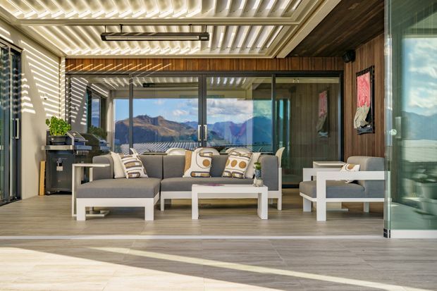 A contemporary outdoor living room or enclosed terrace with a grey sectional sofa, white coffee tables, and matching armchair. The space features timber wall cladding, large glazed sliding doors, a louvred or slatted ceiling, and strong sunlight casting linear shadows across the pale floor. Mountain scenery is visible through the glazing.