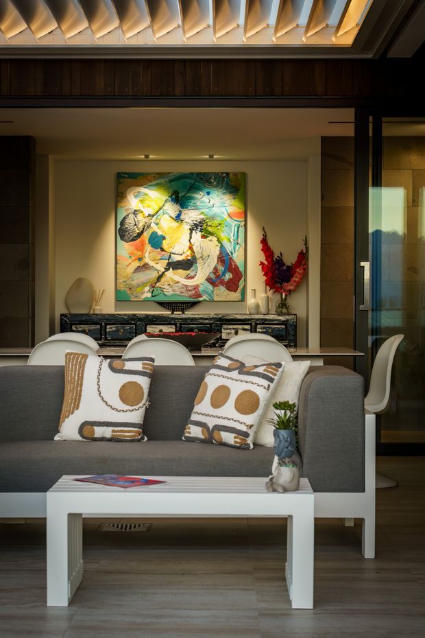 A warmly lit contemporary living and dining area viewed from behind a grey sofa. The room features a white coffee table, patterned decorative cushions, a sculptural console with abstract artwork above it, a dining table with white chairs, dark timber wall panelling, stone-look wall surfaces, and a dramatic illuminated timber ceiling with repeated angled slats. A glazed door or window on the right reflects the exterior landscape.